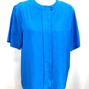 May include: Bright blue short-sleeved blouse. The blouse features a round neckline and a single button closure. The fabric appears to be a lightweight material, with vertical pleats down the front.