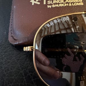May include: Gold-rimmed Ray-Ban sunglasses with mirrored lenses. The lenses reflect a pink and teal pattern. The text "Ray-Ban 50" is visible on the lens. A brown leather case is in the background.