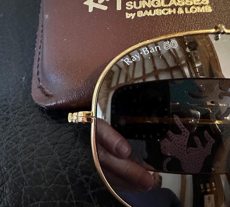 May include: Gold-rimmed Ray-Ban sunglasses with mirrored lenses. The lenses reflect a pink and teal pattern. The text "Ray-Ban 50" is visible on the lens. A brown leather case is in the background.