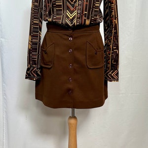 May include: A brown skirt with a button front and two large pockets. The skirt is paired with a long-sleeved shirt with a geometric pattern in brown, orange, and yellow.