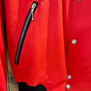 Puede incluir: Primer plano de una chaqueta roja brillante con cremallera negra y herrajes plateados. La chaqueta tiene un pu&ntilde;o acanalado negro y botones plateados. El material parece liso, posiblemente sint&eacute;tico.