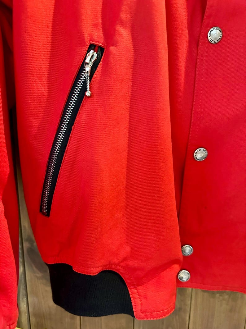 Puede incluir: Primer plano de una chaqueta roja brillante con cremallera negra y herrajes plateados. La chaqueta tiene un pu&ntilde;o acanalado negro y botones plateados. El material parece liso, posiblemente sint&eacute;tico.