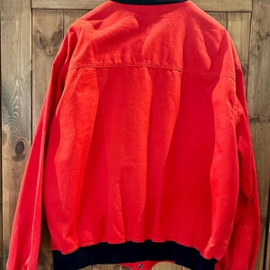 Puede incluir: Una chaqueta roja brillante con cuello, pu&ntilde;os y dobladillo negros. La chaqueta tiene cierre de bot&oacute;n y un estilo cl&aacute;sico de chaqueta bomber. La parte trasera de la chaqueta es visible, mostrando las costuras y los detalles de construcci&oacute;n. La tela es lisa, posiblemente de nailon o poli&eacute;ster.