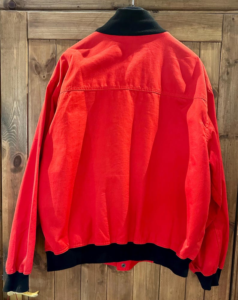 Puede incluir: Una chaqueta roja brillante con cuello, pu&ntilde;os y dobladillo negros. La chaqueta tiene cierre de bot&oacute;n y un estilo cl&aacute;sico de chaqueta bomber. La parte trasera de la chaqueta es visible, mostrando las costuras y los detalles de construcci&oacute;n. La tela es lisa, posiblemente de nailon o poli&eacute;ster.