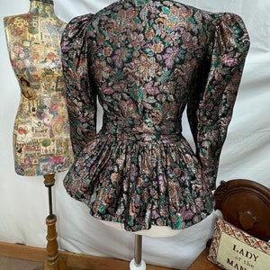 Puede incluir: Una chaqueta de brocado negra, dorada y verde con un dobladillo con volantes y mangas abullonadas. La chaqueta tiene un estampado floral.