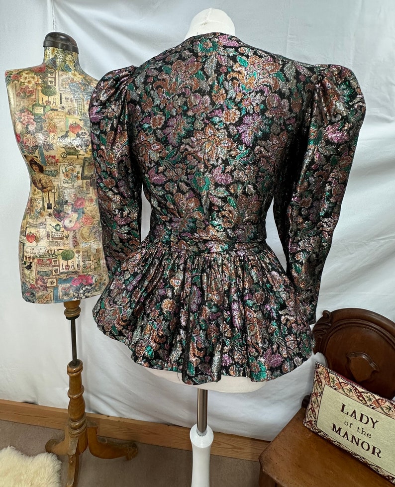 Puede incluir: Una chaqueta de brocado negra, dorada y verde con un dobladillo con volantes y mangas abullonadas. La chaqueta tiene un estampado floral.