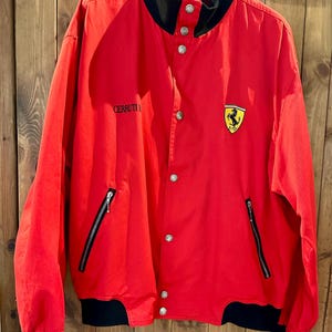Puede incluir: Chaqueta roja con detalles negros, cuello y pu&ntilde;os negros. Cierre de botones, dos bolsillos con cremallera y el logo de Ferrari en el pecho. El texto "CERRUTI" es visible en el pecho izquierdo. Estilo bomber.