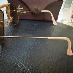 May include: Gold-framed sunglasses with brown lenses. The sunglasses have a classic aviator design with a double bridge and light-coloured ear pieces. A brown case is visible in the background.