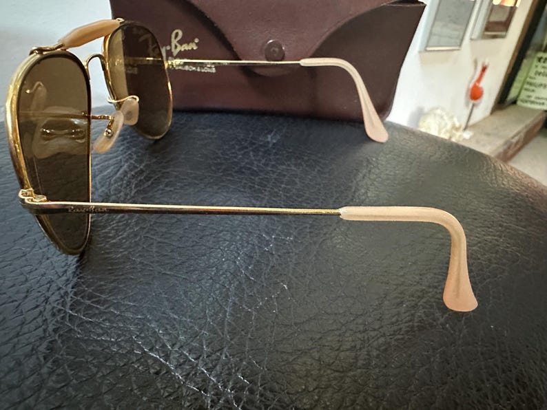 May include: Gold-framed sunglasses with brown lenses. The sunglasses have a classic aviator design with a double bridge and light-coloured ear pieces. A brown case is visible in the background.