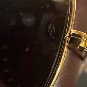 May include: Close-up of gold-toned sunglasses with dark lenses. The frame has a rectangular shape and a hinge detail. The initials "B L" are visible on the lens.