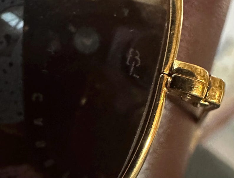 May include: Close-up of gold-toned sunglasses with dark lenses. The frame has a rectangular shape and a hinge detail. The initials "B L" are visible on the lens.