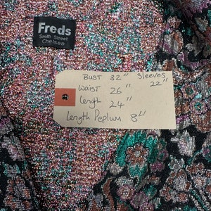 Puede incluir: Una tela met&aacute;lica negra y verde con un patr&oacute;n floral. La tela tiene una etiqueta adjunta con las siguientes medidas: Busto 81 cm, Cintura 66 cm, Largo 61 cm, Largo del Peplum 20 cm, Mangas 56 cm.