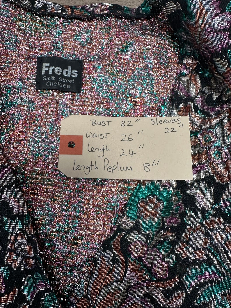 Puede incluir: Una tela met&aacute;lica negra y verde con un patr&oacute;n floral. La tela tiene una etiqueta adjunta con las siguientes medidas: Busto 81 cm, Cintura 66 cm, Largo 61 cm, Largo del Peplum 20 cm, Mangas 56 cm.