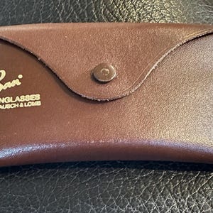 May include: A brown leather sunglasses case with the Ray-Ban logo in gold lettering. The case is rectangular with a curved flap and a button closure. The text "SUNGLASSES by BAUSCH & LOMB" is also visible.