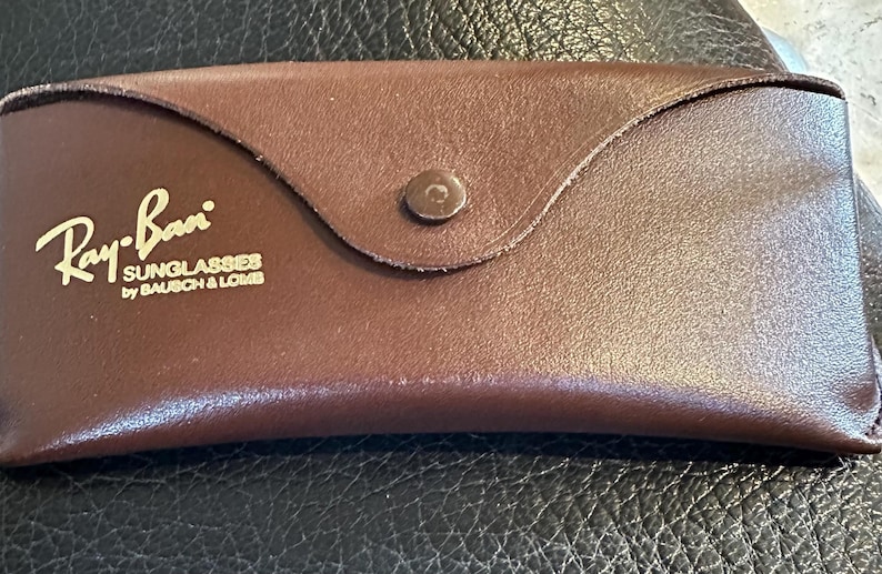 May include: A brown leather sunglasses case with the Ray-Ban logo in gold lettering. The case is rectangular with a curved flap and a button closure. The text "SUNGLASSES by BAUSCH & LOMB" is also visible.