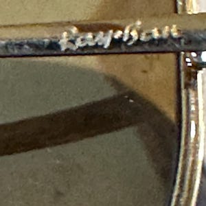 May include: Close-up of sunglasses with the brand name "Ray-Ban" etched on the silver metal frame. The lenses are light brown, and the design is classic and stylish.