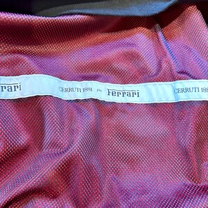 Puede incluir: Primer plano de una tela de malla roja y morada con ribete negro. Una etiqueta azul claro en la parte superior dice "CERRUTI 1881 per Ferrari" en texto negro. La tela tiene un patr&oacute;n texturizado en forma de diamante.