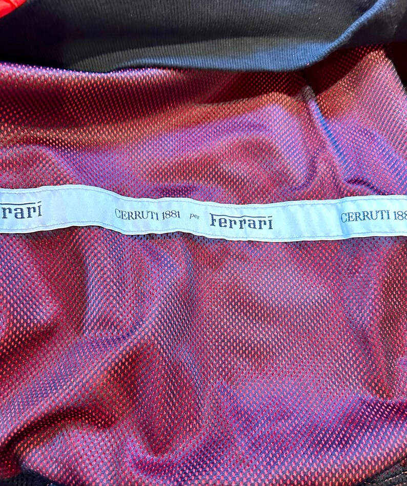 Puede incluir: Primer plano de una tela de malla roja y morada con ribete negro. Una etiqueta azul claro en la parte superior dice "CERRUTI 1881 per Ferrari" en texto negro. La tela tiene un patr&oacute;n texturizado en forma de diamante.