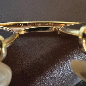 May include: Close-up of gold-toned Ray-Ban sunglasses. The text "B&L RAY-BAN 58[]14" is visible on the frame. The sunglasses have a classic design with a gold frame and dark lenses. The background is a textured brown surface.