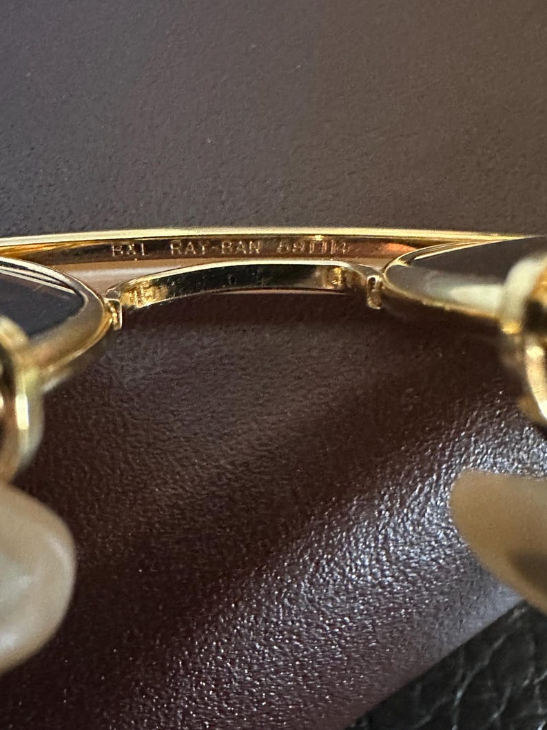 May include: Close-up of gold-toned Ray-Ban sunglasses. The text "B&L RAY-BAN 58[]14" is visible on the frame. The sunglasses have a classic design with a gold frame and dark lenses. The background is a textured brown surface.