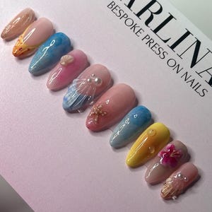 May include: Set of ten press-on nails with a summer beach theme. The nails feature various designs including seashells, starfish, and a flower. The nails are in shades of blue, yellow, and pink.