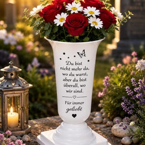 Könnte beinhalten: Eine weiße Grabvase mit schwarzem Schmetterlingsmotiv, Sternen und dem Text: Du bist nicht mehr da, wo du warst, aber du bist überall, wo wir sind. Für immer geliebt. Die Vase ist mit roten Rosen und weißen Margeriten gefüllt.
