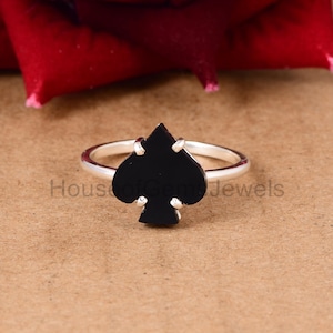 May include: A silver ring with a black spade-shaped stone. The ring is on a brown surface.