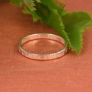 Puede incluir: Un anillo plateado con un diseño repetitivo de medias lunas y líneas. El anillo está sobre una superficie color melocotón, con hojas verdes en el fondo. El anillo es una banda sencilla.