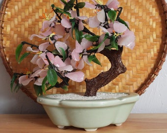 May include: A pink glass flower bonsai tree in a light green ceramic pot. The tree is sitting on a woven bamboo mat.