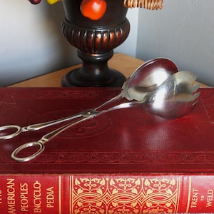 May include: A pair of silver-toned metal salad tongs featuring scissor-style handles with oval finger loops and two spoon-shaped heads, one of which has three tines. The tongs are resting on a red, hardbound book titled The American Peoples Encyclopedia.