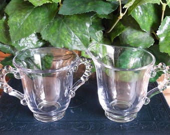 May include: Two clear glass cream and sugar set. The handles are decorated with small, round glass beads. The set is sitting on a dark blue surface, with green leaves in the background.