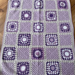 May include: A handmade crochet blanket featuring a patchwork design. The blanket is composed of square motifs in shades of lavender, purple, and white, with a neutral border. The granny square pattern is visible throughout the blanket.