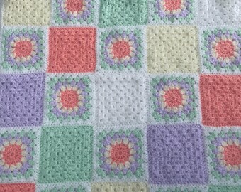 Puede incluir: Una manta de ganchillo hecha a mano con un diseño de patchwork. La manta está compuesta por cuadrados en colores pastel: melocotón, lavanda, amarillo y verde menta. Cada cuadrado está bordeado en blanco, con un motivo floral en el centro.