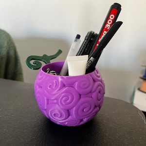 Puede incluir: Un organizador de escritorio esférico morado con un patrón de remolinos. Contiene bolígrafos, rotuladores y un pequeño tubo de crema. El organizador está sobre una superficie oscura.