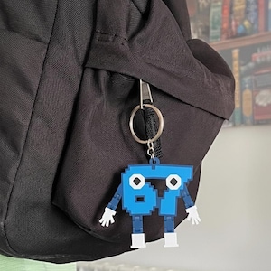 Puede incluir: Un llavero de robot azul pixelado con el número "67" en su cuerpo cuelga de una mochila negra. El robot tiene manos y pies blancos. El llavero está sujeto a un llavero plateado.