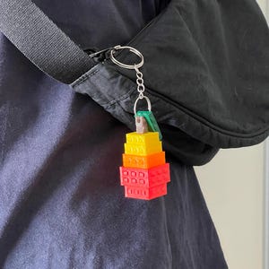 Könnte beinhalten: Ein farbenfroher Schlüsselanhänger mit einem Miniaturstapel aus Bausteinen in Rot, Orange und Gelb. Der Schlüsselanhänger ist an einem silbernen Ring und einem schwarzen Riemen befestigt, was darauf hindeutet, dass er zu einer Tasche oder einem Accessoire gehört.