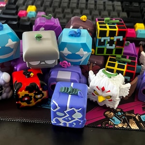 Puede incluir: Una colección de keycaps artesanales coloridos con varios diseños, incluyendo formas geométricas, personajes de dibujos animados y patrones abstractos. Los keycaps son en tonos de azul, morado, rojo, amarillo y blanco. Los keycaps están sobre una superficie estampada.