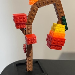 May include: A 3D-printed sculpture of a tree made of blocks. The trunk and branches are brown, with red, orange, and yellow blocks representing the leaves. The sculpture sits in a black square base.