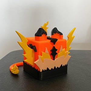 Könnte beinhalten: Eine 3D-gedruckte, würfelförmige Skulptur in Orange, Schwarz und Gelb. Der Würfel weist stilisierte Flammen- und Blitzmuster auf. Ein kleiner oranger Schwanz erstreckt sich von der Basis. Die Skulptur steht auf einer schwarzen Oberfläche.