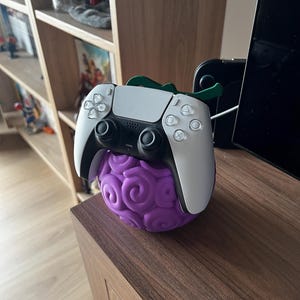 May include: A white and black PlayStation 5 controller sits on a purple, textured fruit-shaped stand with green detailing. The stand features a swirling pattern and is designed to hold the controller.