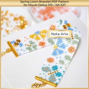 Op de afbeelding: Een kralenarmband met een bloemenpatroon in blauw, groen, oranje en geel. De armband is wit met goudkleurige sluitingen en ketting. De tekst "Nota Arta" is zichtbaar.