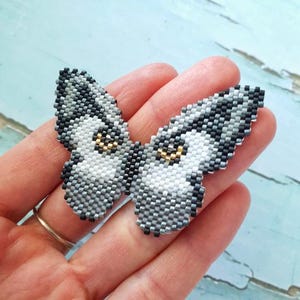 K&ouml;nnte beinhalten: Eine Brosche in Form eines Schmetterlings aus grauen, wei&szlig;en und schwarzen Perlen. Der Schmetterling hat goldene Akzente f&uuml;r die Augen.