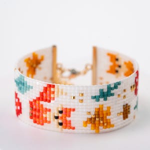 Puede incluir: Pulsera de cuentas blanca con un patr&oacute;n colorido de zorros, &aacute;rboles y otras formas. La pulsera presenta cuentas rojas, naranjas, turquesas y doradas. El cierre es de color dorado.