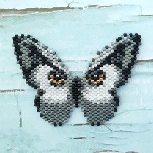 K&ouml;nnte beinhalten: Ein Perlen-Schmetterling mit grauen, wei&szlig;en und schwarzen Perlen. Der Schmetterling hat goldene Augen.