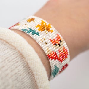 Puede incluir: Una pulsera de cuentas con fondo blanco y motivos florales coloridos. La pulsera presenta cuentas naranjas, rojas, turquesas y doradas. La pulsera mide aproximadamente 3,8 cm de ancho.