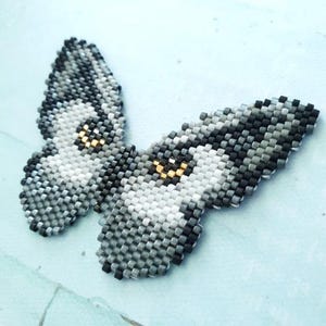 K&ouml;nnte beinhalten: Ein grauer und wei&szlig;er Perlen-Schmetterling mit goldenen Akzenten. Der Schmetterling besteht aus kleinen, quadratischen Perlen und hat ein detailliertes Design.