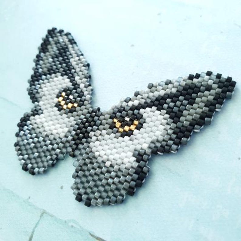 K&ouml;nnte beinhalten: Ein grauer und wei&szlig;er Perlen-Schmetterling mit goldenen Akzenten. Der Schmetterling besteht aus kleinen, quadratischen Perlen und hat ein detailliertes Design.