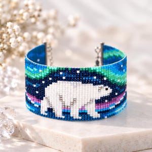 May include: A wide cuff bracelet crafted from small glass beads, featuring a white polar bear centered against a dark blue night sky. The design includes vibrant green and purple bands representing the aurora borealis above and below.