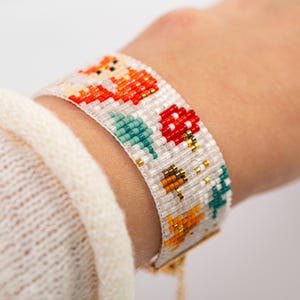 Puede incluir: Pulsera de cuentas con un dise&ntilde;o tem&aacute;tico de la naturaleza. La pulsera presenta un fondo blanco con zorros naranjas, setas rojas y &aacute;rboles turquesas. La pulsera mide aproximadamente 3,8 cm de ancho y tiene un cierre dorado.