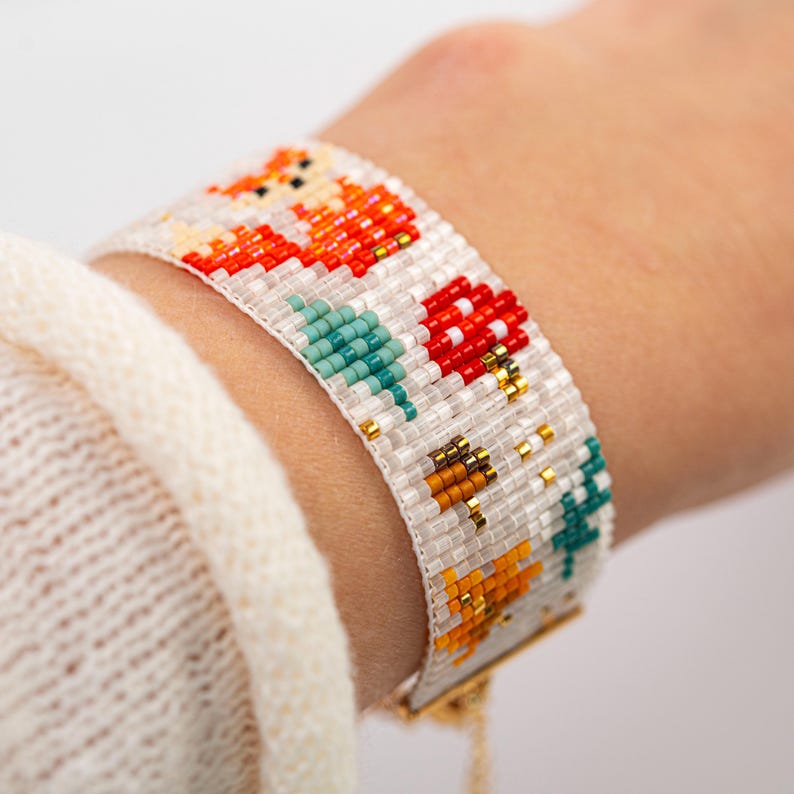 Puede incluir: Pulsera de cuentas con un dise&ntilde;o tem&aacute;tico de la naturaleza. La pulsera presenta un fondo blanco con zorros naranjas, setas rojas y &aacute;rboles turquesas. La pulsera mide aproximadamente 3,8 cm de ancho y tiene un cierre dorado.